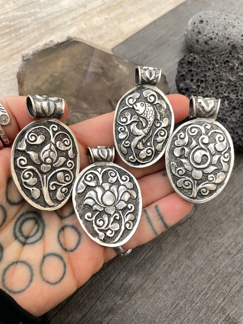 May include: Four silver pendants featuring intricate floral and fish designs. The pendants are oval shaped and have a detailed, textured surface.