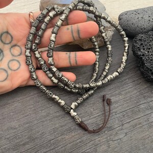 May include: A silver skull bead necklace with a brown cord closure. The necklace is made of small, round beads with a skull design.