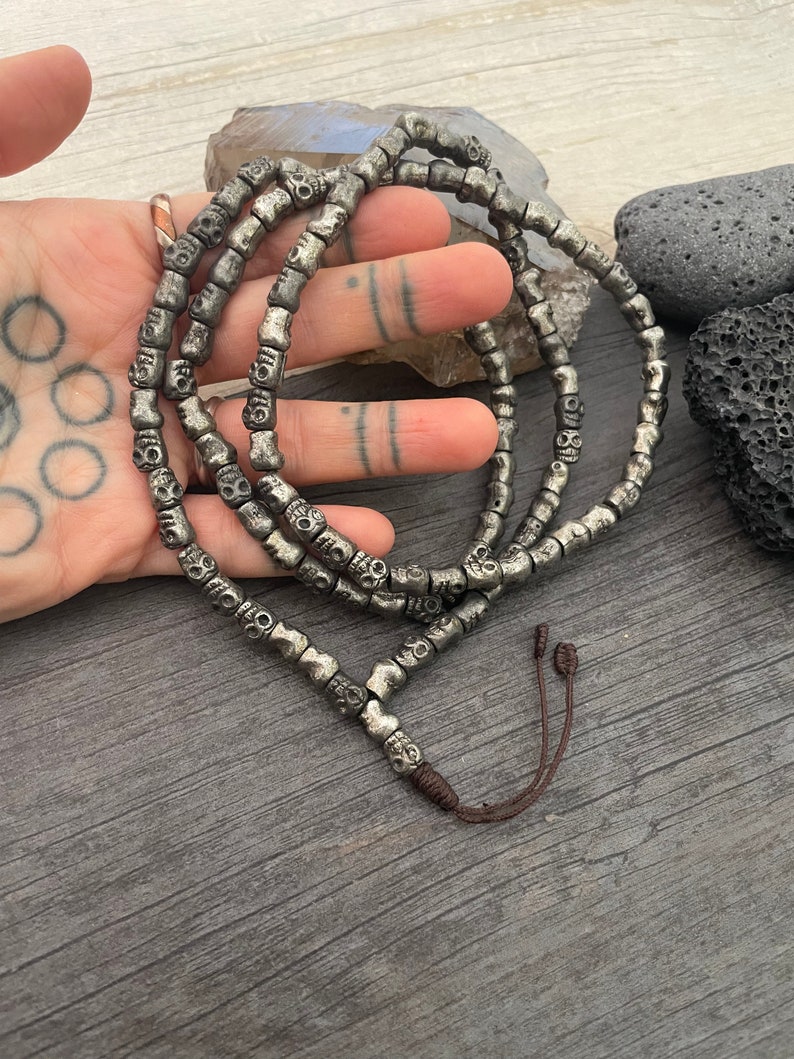 May include: A silver skull bead necklace with a brown cord closure. The necklace is made of small, round beads with a skull design.