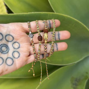 May include: A set of four thin bracelets made from multicolored braided cord. Each bracelet features a unique charm, including a silver-toned eternal knot, a brown rudraksha seed, and a gold-toned Buddha head, all accented with small metallic beads.