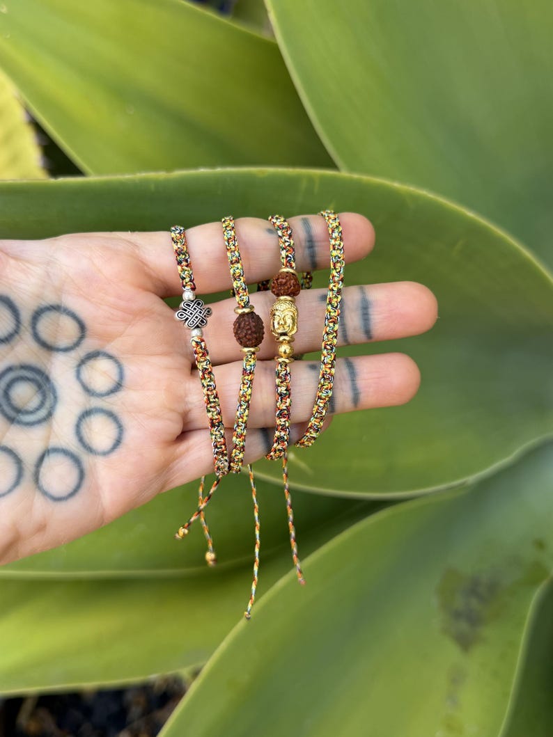 May include: A set of four thin bracelets made from multicolored braided cord. Each bracelet features a unique charm, including a silver-toned eternal knot, a brown rudraksha seed, and a gold-toned Buddha head, all accented with small metallic beads.