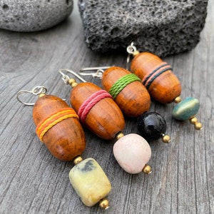 May include: Four handmade earrings with wooden barrel beads wrapped in colorful thread. Each earring has a different gemstone or bead, including a pale yellow stone, a pink stone, a black bead, and a blue-green bead. The earrings are on a gray wooden surface.