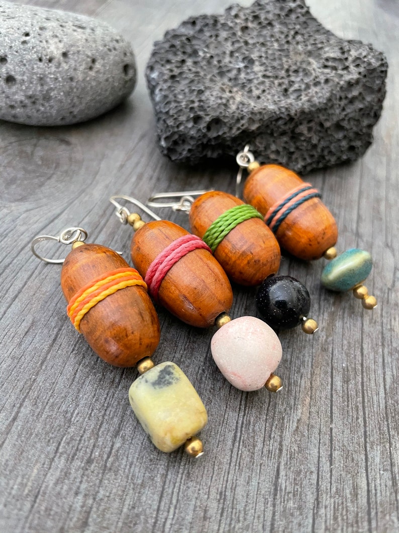 May include: Four handmade earrings with wooden barrel beads wrapped in colorful thread. Each earring has a different gemstone or bead, including a pale yellow stone, a pink stone, a black bead, and a blue-green bead. The earrings are on a gray wooden surface.