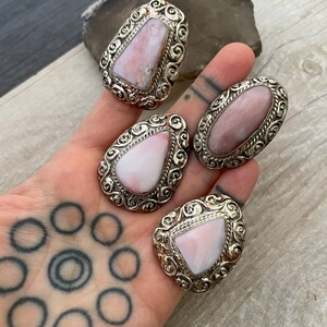 May include: Four silver rings with pink opal stones set in ornate silver settings. The rings are displayed on a hand with a tattoo.