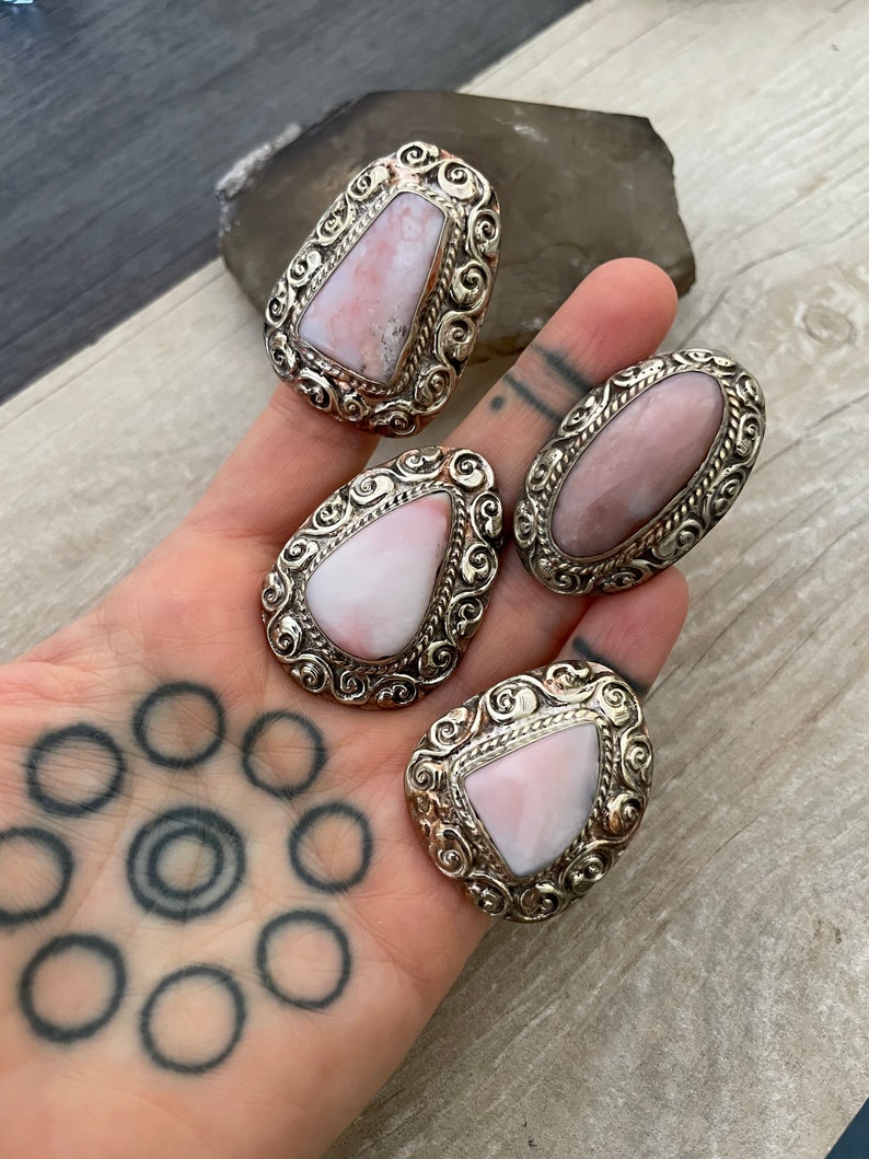 May include: Four silver rings with pink opal stones set in ornate silver settings. The rings are displayed on a hand with a tattoo.