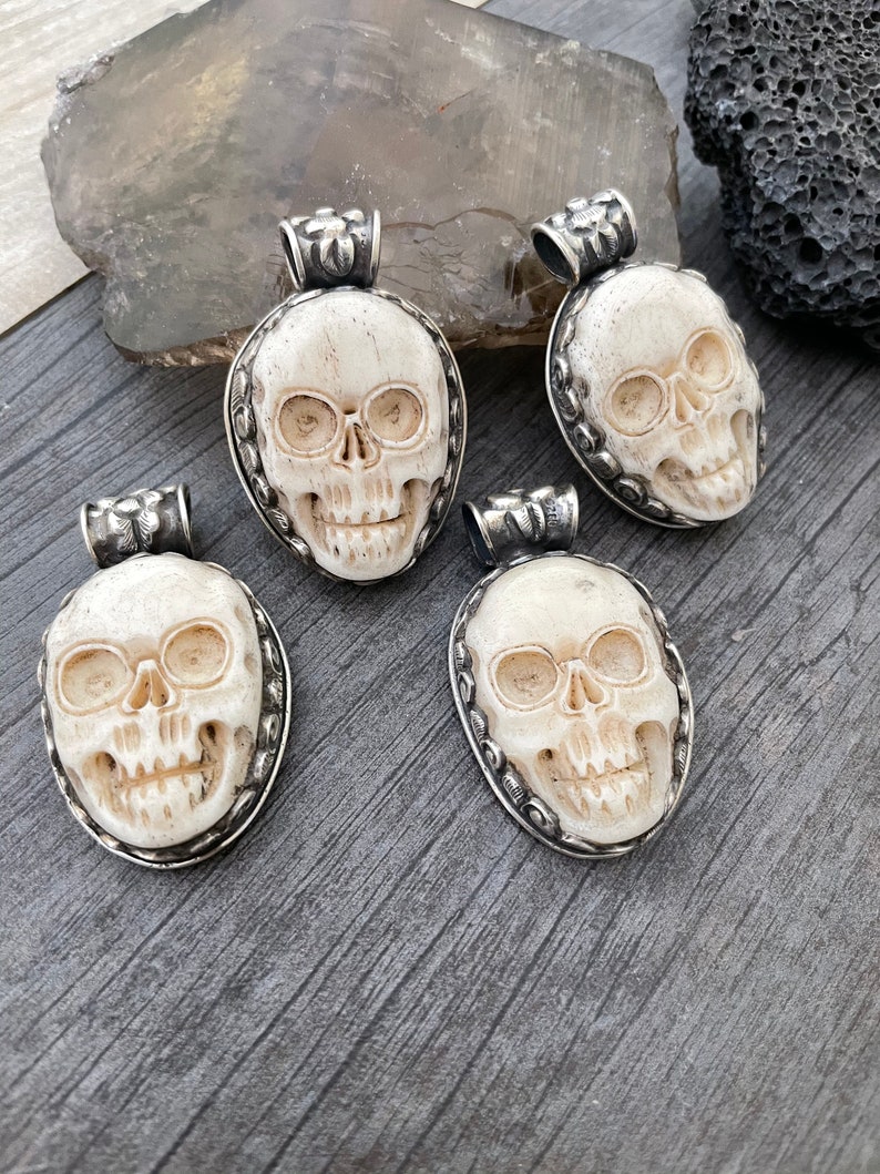 May include: Four white bone skull pendants with silver trim. Each pendant features a detailed skull design with eye sockets, a nose, and a mouth.