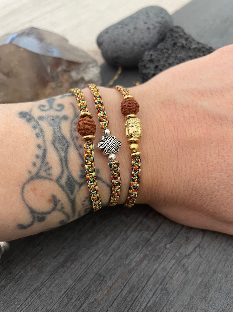 May include: Three braided bracelets made of multicolored yellow, red, and green cord. One bracelet features a brown rudraksha bead, another has a silver-toned eternal knot charm, and the third includes a gold-toned Buddha head and a rudraksha bead.