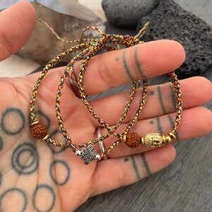 May include: Two braided bracelets made of multicolored cord in shades of yellow, orange, and black. One bracelet features a silver-toned eternal knot charm, while the other includes a gold-toned Buddha head charm and brown textured beads.