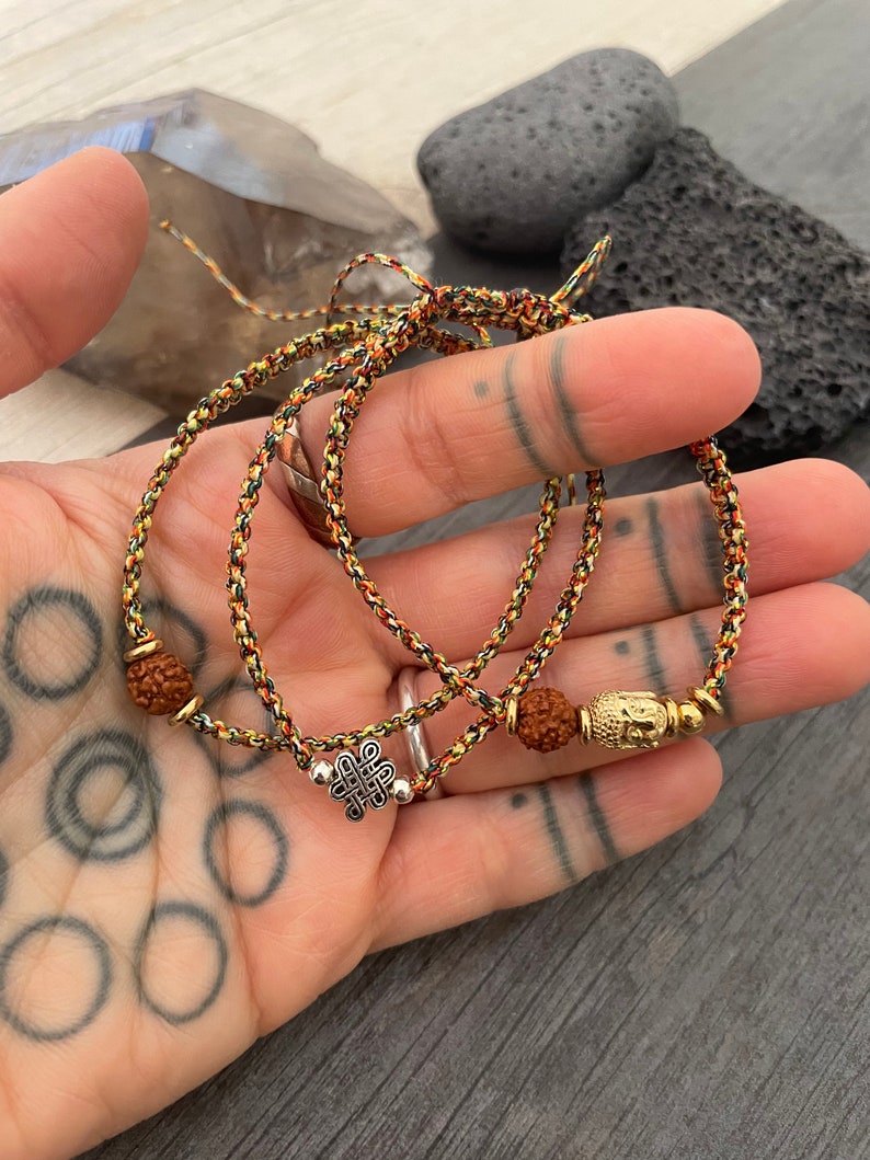 May include: Two braided bracelets made of multicolored cord in shades of yellow, orange, and black. One bracelet features a silver-toned eternal knot charm, while the other includes a gold-toned Buddha head charm and brown textured beads.