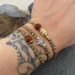 May include: Three braided bracelets made of multicolored thread in shades of yellow, green, and red. One bracelet features a brown textured bead and a gold-toned Buddha charm. Another includes a silver-toned eternal knot charm and a brown bead.