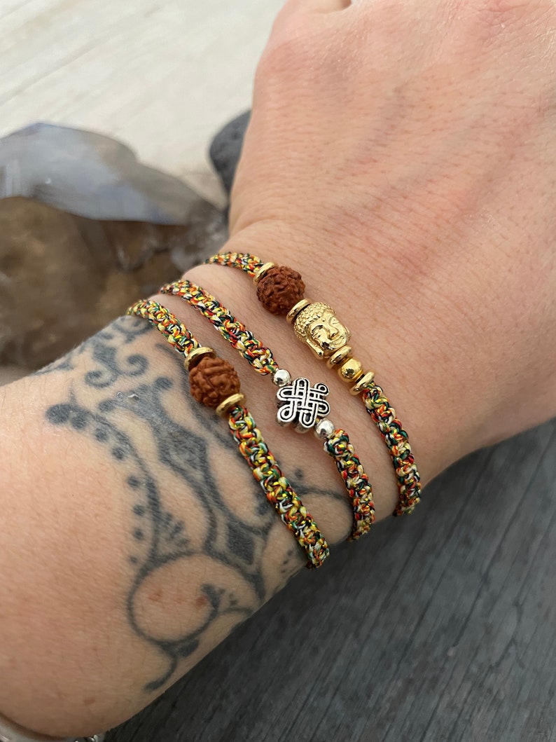 May include: Three braided bracelets made of multicolored thread in shades of yellow, green, and red. One bracelet features a brown textured bead and a gold-toned Buddha charm. Another includes a silver-toned eternal knot charm and a brown bead.