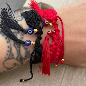 Puede incluir: Primer plano de tres pulseras hechas a mano. Dos pulseras negras presentan cuentas azules de ojo turco y detalles dorados. La tercera pulsera es roja con un diseño en forma de corazón, borlas rojas y cuentas rojas de ojo turco. Todas las pulseras tienen cordones ajustables.