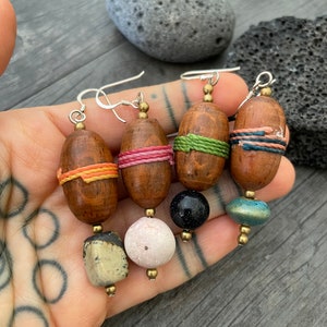 May include: Four pairs of handcrafted earrings with wooden, oval-shaped beads wrapped in colorful thread. Each earring features a unique combination of beads, including stone, pink, black, and teal, suspended from silver hooks.