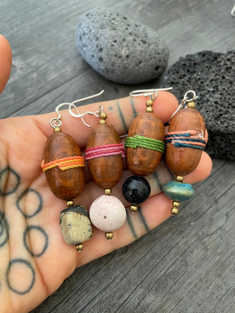 May include: Four pairs of handcrafted earrings with wooden, oval-shaped beads wrapped in colorful thread. Each earring features a unique combination of beads, including stone, pink, black, and teal, suspended from silver hooks.