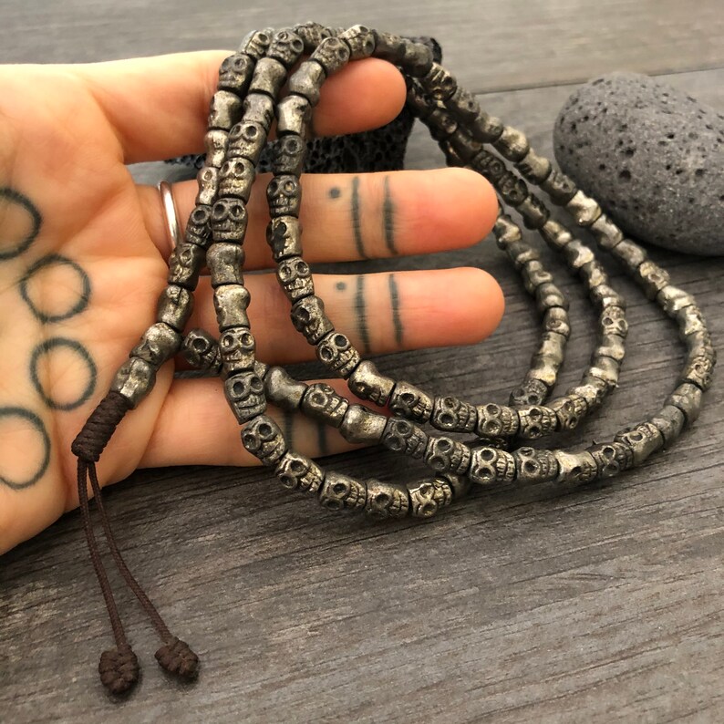 May include: A silver skull bead mala necklace with a brown cord tassel. The necklace is made of multiple strands of beads and is laid out on a wooden surface.