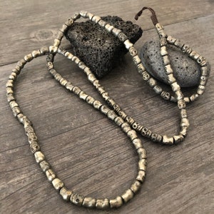 May include: A silver skull bead necklace with a brown cord. The necklace is made of many small skull beads and is laid out on a wooden surface.