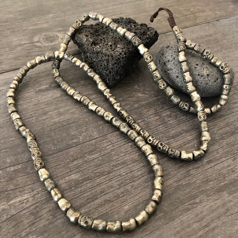 May include: A silver skull bead necklace with a brown cord. The necklace is made of many small skull beads and is laid out on a wooden surface.