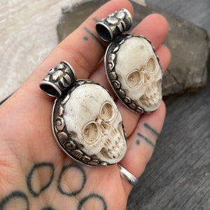 May include: Two white bone skull pendants with silver accents. The skulls are carved with intricate details and have a floral design around the edges. The pendants are attached to silver chains.