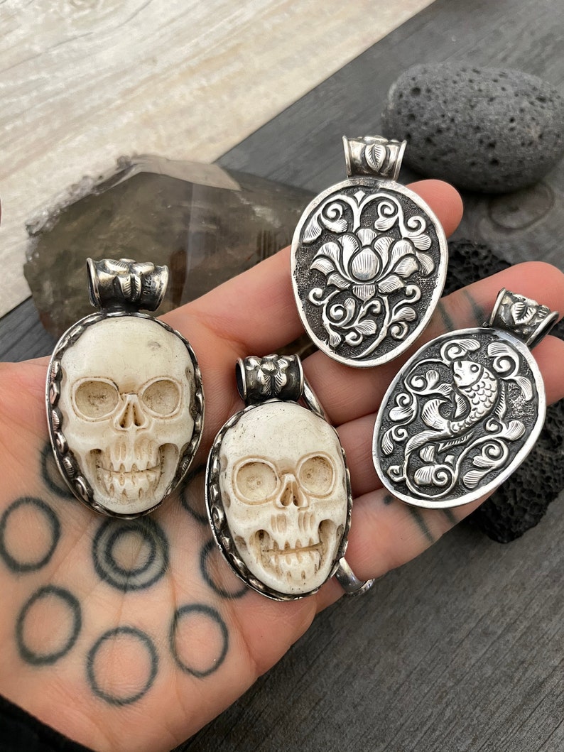 May include: Four silver pendants with intricate designs. Two pendants feature a skull design, while the other two feature floral and fish designs.