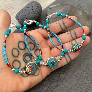 Puede incluir: Tres pulseras de cuentas turquesa y plata con dijes redondos decorativos. Los dijes son de metal y tienen detalles en turquesa, rojo y azul.
