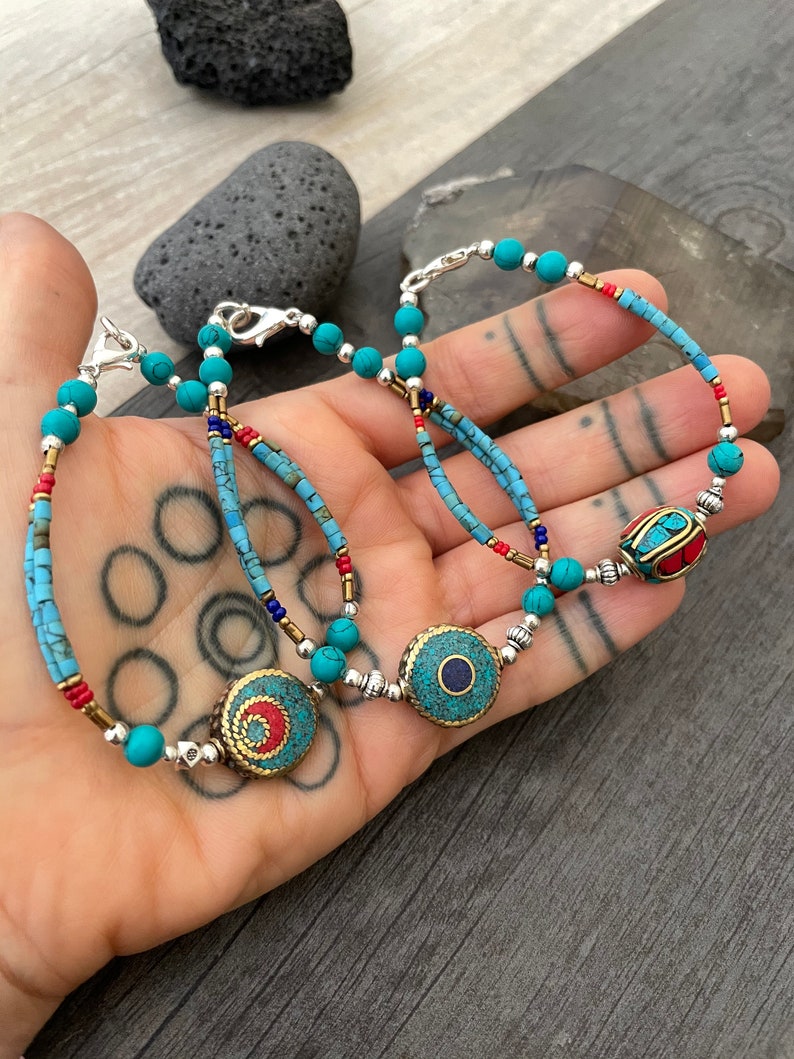 Puede incluir: Tres pulseras de cuentas turquesa y plata con dijes redondos decorativos. Los dijes son de metal y tienen detalles en turquesa, rojo y azul.