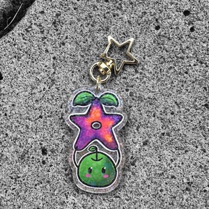May include: A green and purple star-shaped charm with a cute face and a green plant growing from the top. The charm is attached to a gold star-shaped keychain.