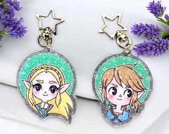 May include: Two round keychains with gold star-shaped charms. Each keychain features a cartoon character with large eyes and blonde hair. One character is wearing a blue tunic and the other is wearing a green tunic.