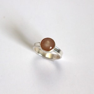 Puede incluir: Un anillo de plata con una piedra redonda de color marrón claro engastada en el centro.