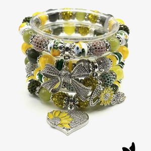May include: Stack of beaded bracelets in shades of yellow, green, and silver. The bracelets feature various bead styles, including round, faceted, and decorative beads. Some bracelets have bow and sunflower charms, and a heart-shaped pendant.