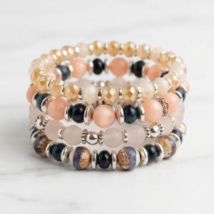 Peut inclure: Empilement de quatre bracelets de perles aux tons pêche, crème, noir et bleu. Les bracelets présentent une variété de types de perles, dont des perles à facettes, rondes et à motifs, séparées par des entretoises argentées. Les bracelets sont empilés sur une surface en marbre blanc.