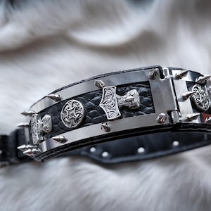 Peut inclure: Un collier en cuir noir avec des détails en métal argenté et des pointes. Le collier présente des plaques décoratives en métal avec des motifs complexes, dont un marteau et des motifs circulaires. Il possède une boucle et un anneau en D.