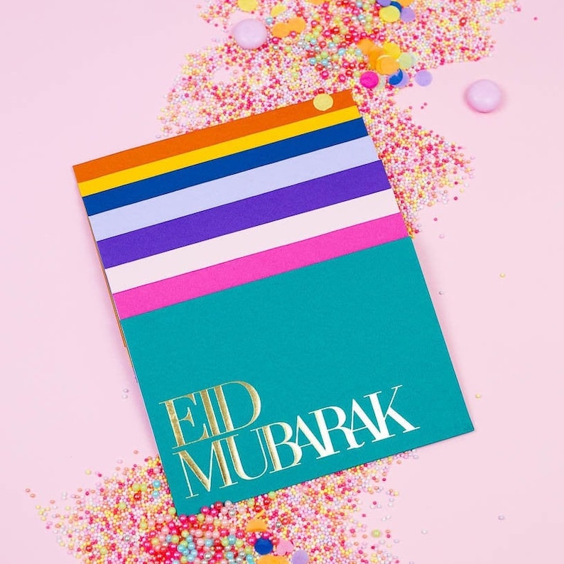 Może przedstawiać: Turkusowa kartka z pozłacanymi literami, na kt&oacute;rej widnieje napis "Eid Mubarak". Kartka jest złożona z warstw kolorowych pask&oacute;w w kolorze r&oacute;żowym, białym, fioletowym, niebieskim, pomarańczowym i ż&oacute;łtym.