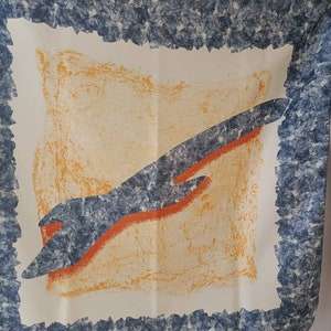May include: A square scarf with a white background, featuring a central design of an orange and blue abstract shape. The scarf has a textured blue border with a jagged edge, and an orange and yellow center.
