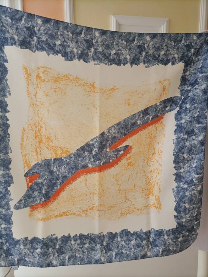 May include: A square scarf with a white background, featuring a central design of an orange and blue abstract shape. The scarf has a textured blue border with a jagged edge, and an orange and yellow center.