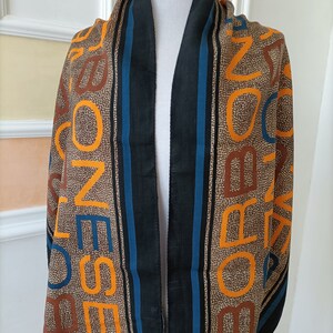 May include: A brown scarf with a repeating pattern of orange and brown letters. The scarf has a black border with blue stripes. The scarf is draped over a white mannequin.