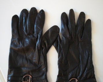 Könnte beinhalten: Ein Paar schwarze Lederhandschuhe. Jeder Handschuh hat einen dekorativen Riemen mit einer silberfarbenen Schnalle in der Nähe des Handgelenks. Die Handschuhe sind aus weichem, strukturiertem Leder gefertigt.