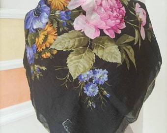 May include: A square black scarf with a vibrant floral print. The design features large pink peonies, blue and yellow flowers, and green leaves. The scarf has a green border and a signature in the corner.