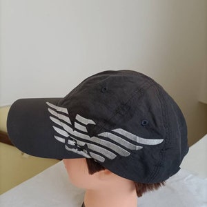 May include: A dark gray baseball cap with a silver eagle design on the side. The cap has a curved brim and a fabric strap for adjustment. The cap is displayed on a mannequin head.