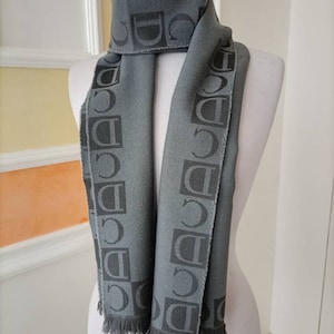 May include: A gray scarf with a repeating pattern of the letters "D" and "C" in a darker shade of gray. The scarf has fringed edges and is draped over a mannequin. The scarf is made of a soft material.