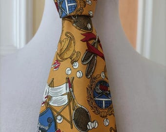 May include: Mustard yellow tie with a repeating pattern of tennis-related imagery, including tennis rackets, tennis balls, trophies, and a tennis court. The tie features a classic design with a pointed end and is displayed on a white mannequin.