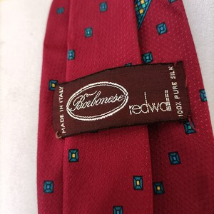 May include: A red silk tie with a repeating pattern of teal and yellow squares. The tie has a textured surface and a label that reads "Barbanese redwall 100% Pure Silk Made in Italy."