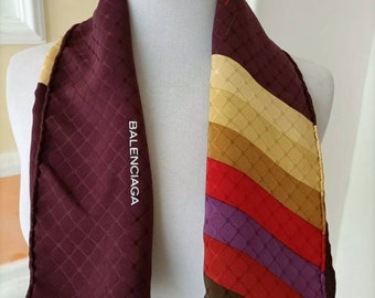 Könnte beinhalten: Burgunderfarbener Schal mit Rautenmuster und dem Wort "BALENCIAGA" in Weiß. Der Schal hat einen Abschnitt mit diagonalen Streifen in Gelb-, Rot-, Lila- und Brauntönen. Er ist über einer weißen Schaufensterpuppe drapiert.