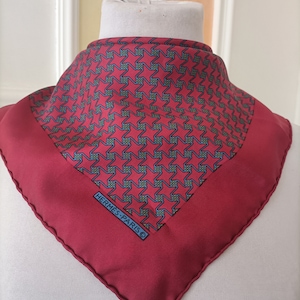 May include: A square, burgundy silk scarf with a repeating star pattern in teal and dark blue. The scarf has the text "HERMES-PARIS" in a dark blue rectangle in the corner. The scarf is displayed on a white mannequin head.