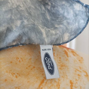 May include: A silk scarf with a blue and white marbled pattern. The scarf has a tag with the Ford logo and the text "100% Seta". The background has an orange and yellow pattern.