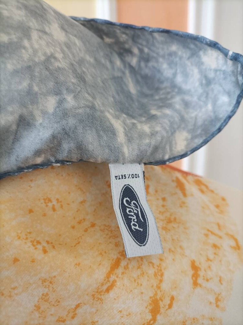May include: A silk scarf with a blue and white marbled pattern. The scarf has a tag with the Ford logo and the text "100% Seta". The background has an orange and yellow pattern.