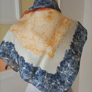 May include: A square scarf with a unique design. The scarf features a white background with orange and yellow brushstrokes, a blue and white textured border, and a blue and white patterned top.