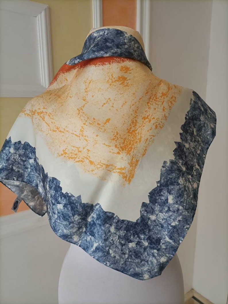 May include: A square scarf with a unique design. The scarf features a white background with orange and yellow brushstrokes, a blue and white textured border, and a blue and white patterned top.