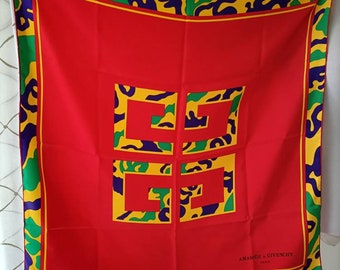 Könnte beinhalten: Ein quadratisches rotes Tuch mit einem gelb, grün und lila gemusterten Rand. Die Mitte zeigt ein großes gelbes und grünes geometrisches Design. Der Text "AMARIGE + GIVENCHY PARIS" ist unten rechts aufgedruckt.