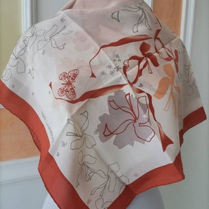 Può includere: Una sciarpa quadrata di seta con un design floreale e a farfalla. La sciarpa presenta uno sfondo color crema con bordi e accenti color ruggine. Il testo "TED LAPIDUS" è visibile sulla sciarpa.