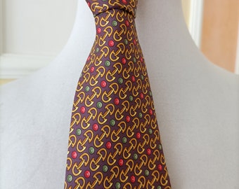 May include: A patterned necktie with a repeating design of gold-colored shapes, red circles, and green circles on a dark purple background. The tie is knotted and displayed on a white mannequin head. The tie is made of a smooth, possibly silk, material.